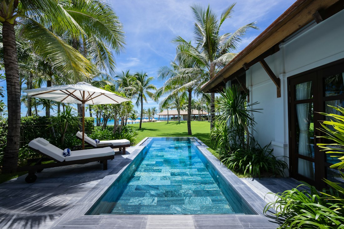 Villa tropicale blanche au toit de bois, piscine étroite bordée de transats, cocotiers et pelouse.