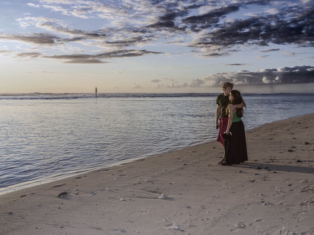 Couple enlacé au bord du lagon sur une plage de sable clair, sous un ciel nuageux de fin de journée, elle en jupe longue noire et haut vert uni, lui en t-shirt sombre