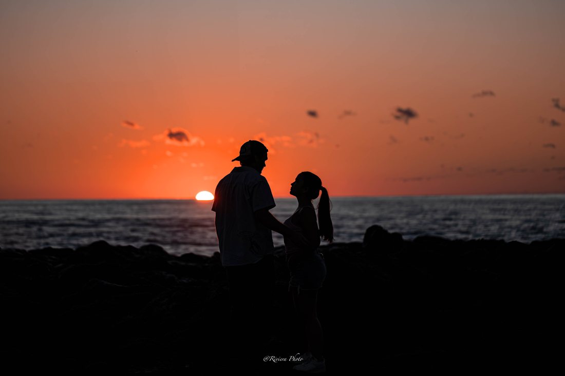 Silhouette d'un couple face à face sur des rochers, devant l'océan, au moment où le soleil touche l'horizon dans un ciel orange.
