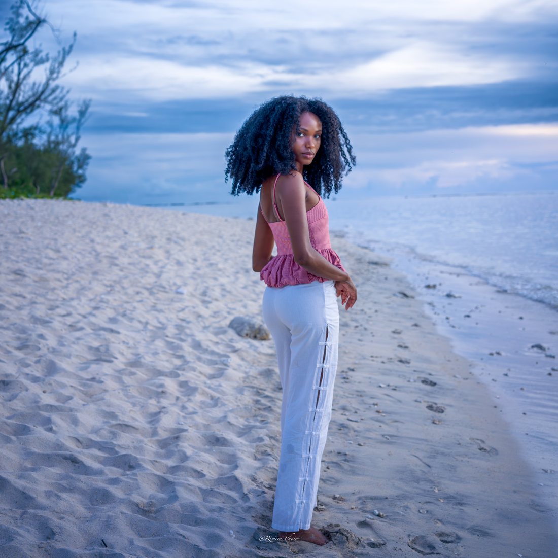 Jeune femme de dos, tournée vers l'objectif, en haut rose à petits carreaux et pantalon blanc, pieds nus sur le sable clair au bord du lagon, filaos à gauche, lumière bleutée de fin de journée