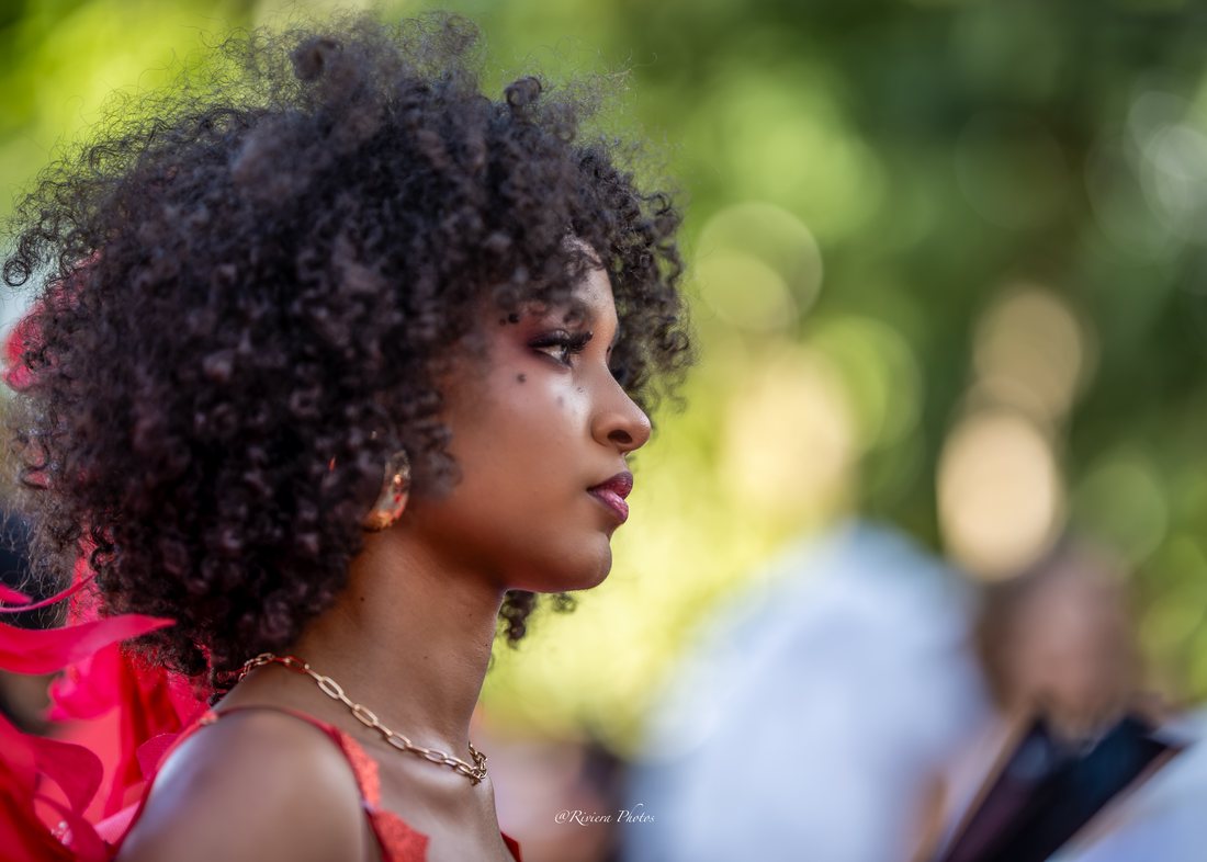 Portrait de profil d'une jeune femme aux cheveux bouclés, robe rouge et collier doré, devant un fond de verdure flou.