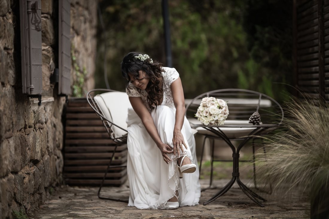 Pendant les préparatifs, la mariée en robe blanche attache sa chaussure, assise sur une terrasse de pierre, son bouquet de roses posé sur la table à côté d'elle.