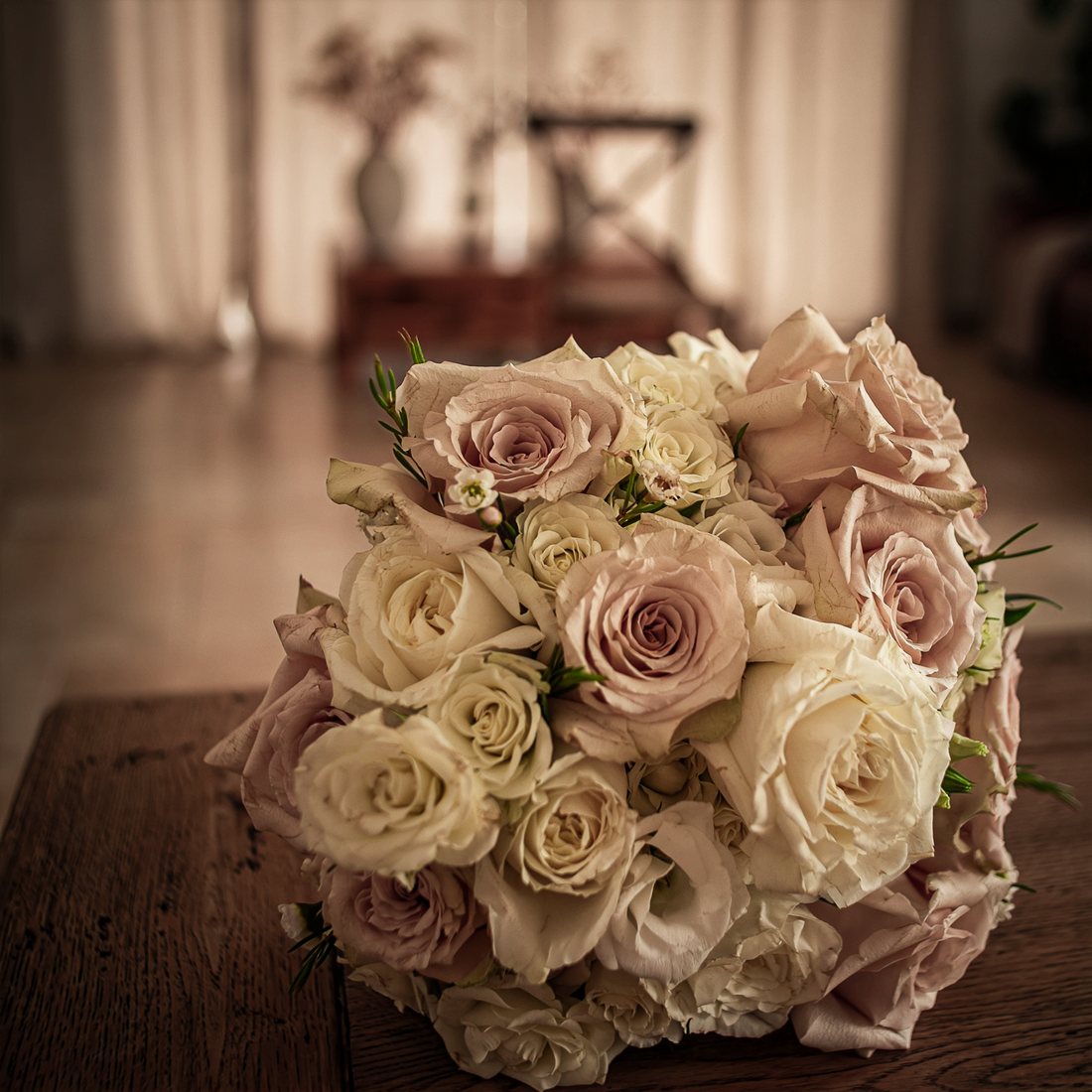 Bouquet de mariée en roses blanches et rose poudré, posé sur une table en bois, dans une pièce aux rideaux clairs.
