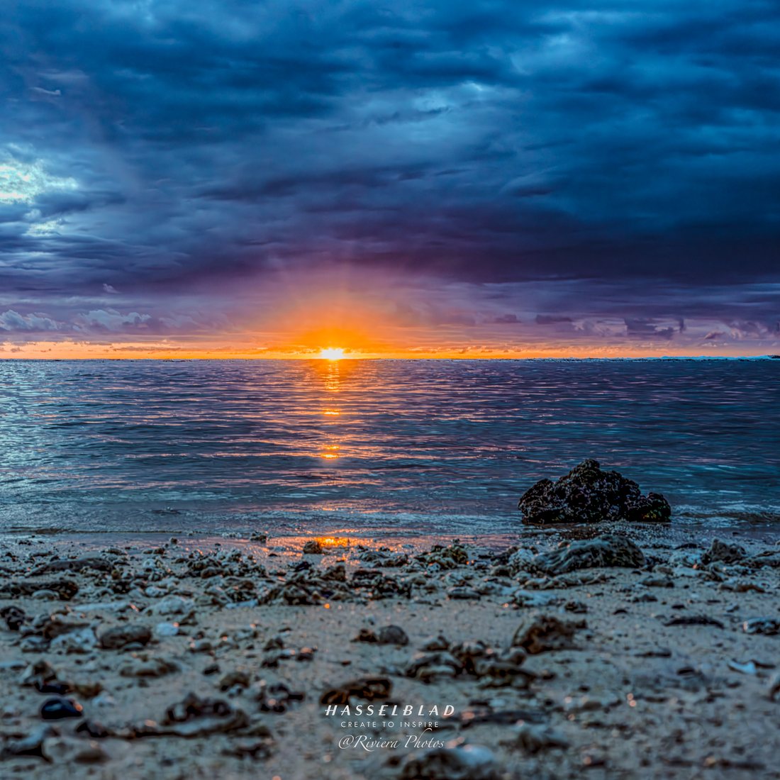 Soleil couchant sur la mer, plage de galets et de corail au premier plan, ciel d'orage bleu.